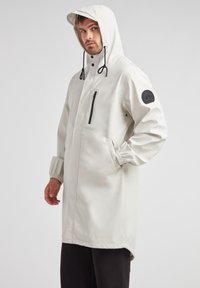Giacca bianca impermeabile con cappuccio, tasca con zip nera, polsini elasticizzati e patch con logo sul braccio sinistro. Design a texture liscia, lunghezza al ginocchio.