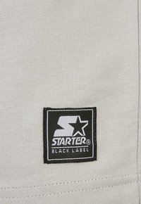 Grått stoff med ribbet tekstur som har et svart og hvitt rektangulært logopatch som leser "STARTER BLACK LABEL."