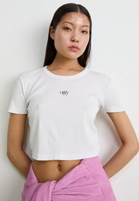 Cropped top bianco a coste con collo tondo, maniche corte e un piccolo logo nero sul petto. Indossato con una gonna avvolgente rosa testurizzata.