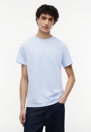 Νεαρός άνδρας με κατσαρά μαλλιά, που φοράει ανοιχτό μπλε κοντομάνικο μπλουζάκι και σκούρο τζιν, στέκεται με το ένα χέρι στην τσέπη μπροστά σε λευκό φόντο.