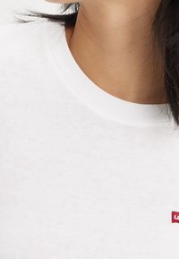 Valkoinen puuvillainen t-paita, jossa on ribbbattu pyöreä kaula-aukko ja pieni punainen brodeerattu logo vasemmalla puolella. Sileä pinta.