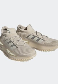 Beige urheilusneakerit, joissa on neulottu yläosa, joustava nilkkatuki, harmaat raidat ja teksturoitu pohja pidon parantamiseksi.