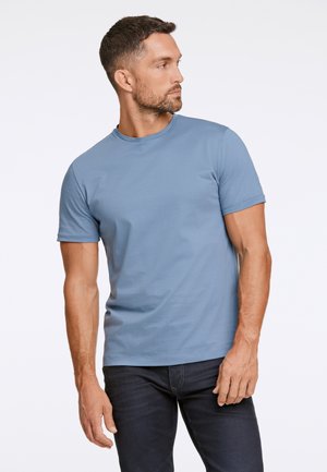 Niebieska koszulka z krótkim rękawem, uszyta z miękkiego materiału, z okrągłym dekoltem i dopasowanym krojem, noszona z ciemnymi dżinsami.