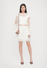 Λευκό φόρεμα από δαντέλα με μακριά διάφανα μανίκια, στρογγυλή λαιμόκοψη και κομμένο σχέδιο στη μέση. Φοριέται με καφέ ψηλές γόβες.