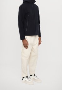 Námořnická kapucí jacket s předním zipem a bočními kapsami, spárovaná s krémovými texturovanými kalhoty a bílými teniskami s černými akcenty.