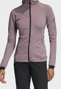 Chaqueta ajustada de cremallera en tejido texturizado de color púrpura claro, con mangas largas, cuello alto y detalles en cremallera negra. Incluye bolsillos laterales.