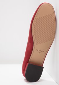 Chaussure plate en daim rouge avec un bout arrondi et un petit talon carré. La semelle présente un motif texturé pour l'adhérence et porte l'impression de la marque.