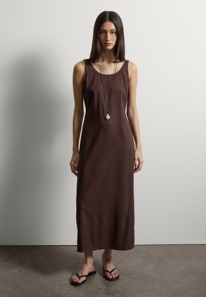 Femme debout contre un mur uni portant une robe maxi marron sans manches, un long collier pendentif et des sandales noires à bout carré.