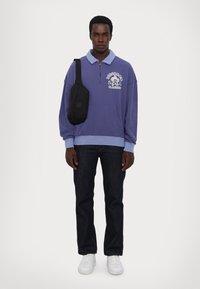 Fialový sveter s svetlomodrým golierom a bielym grafickým potlačou, skombinovaný s tmavými džínsami a bielymi teniskami. Čierna crossbody taška.