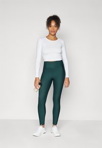Top corto de manga larga blanco combinado con leggings de tiro alto de color verde oscuro. La modelo sostiene una botella de agua de color claro y lleva zapatillas deportivas blancas.