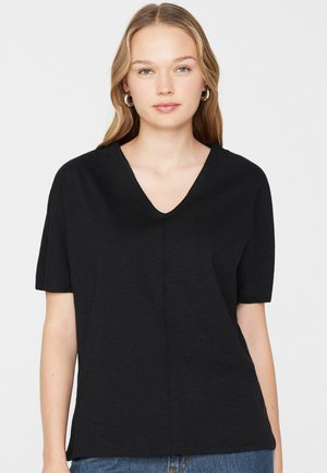 Giovane donna con capelli castano chiaro che indossa orecchini a cerchio d'argento, una maglietta nera a maniche corte con scollo a V e jeans blu su uno sfondo bianco.