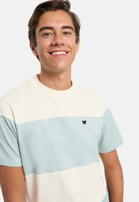 Camiseta de manga corta con rayas horizontales en crema y azul claro, que presenta un pequeño logo en navy en el pecho. Tela de algodón suave.