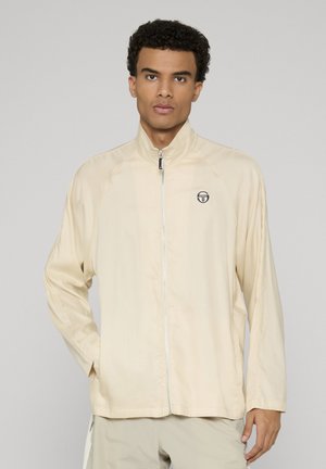 Giovane uomo che indossa una giacca beige con zip e collo alto, in piedi con una mano in tasca contro uno sfondo chiaro e uniforme.