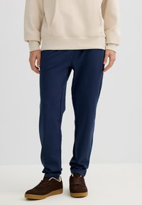 Persona con pantalones casuales azul marino, sudadera beige y zapatillas marrones, de pie frente a un fondo claro y liso.