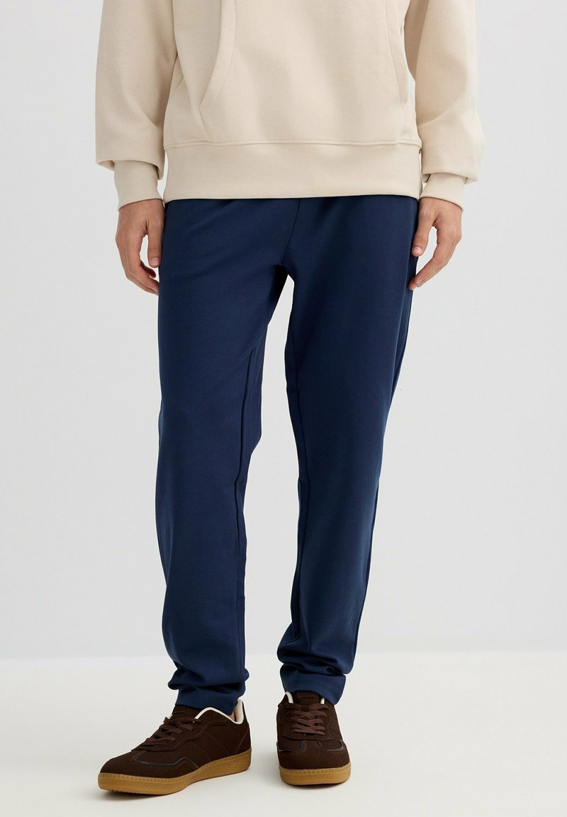 Persona con pantalones casuales azul marino, sudadera beige y zapatillas marrones, de pie frente a un fondo claro y liso.