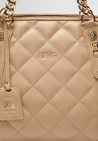 Borsa a tracolla in pelle beige trapuntata con hardware in tonalità oro, con il nome del marchio 'LIUJO' in rilievo e un'etichetta staccabile con la dicitura 'Kate RE-EDITION 2022'.