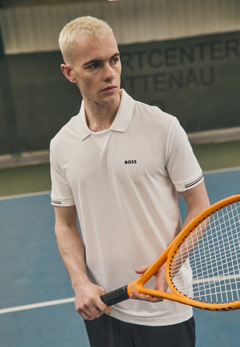 Λευκό μπλουζάκι πόλο με μικρό μαύρο λογότυπο, μαύρες λωρίδες στις μανίκες, φορεμένο με μαύρες σορτς. Το άτομο κρατά μια πορτοκαλί ρακέτα τένις.