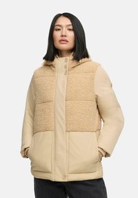 Beige pufferjack met een tan fleece paneel aan de voorkant, voorzien van een capuchon, drukknoopsluiting en zijzakken. Soepele textuur en oversized pasvorm.