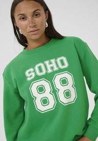 Zelená mikina s potiskem "SOHO 88" v tučných bílých písmenech. Má kulatý výstřih a měkký, texturovaný povrch.