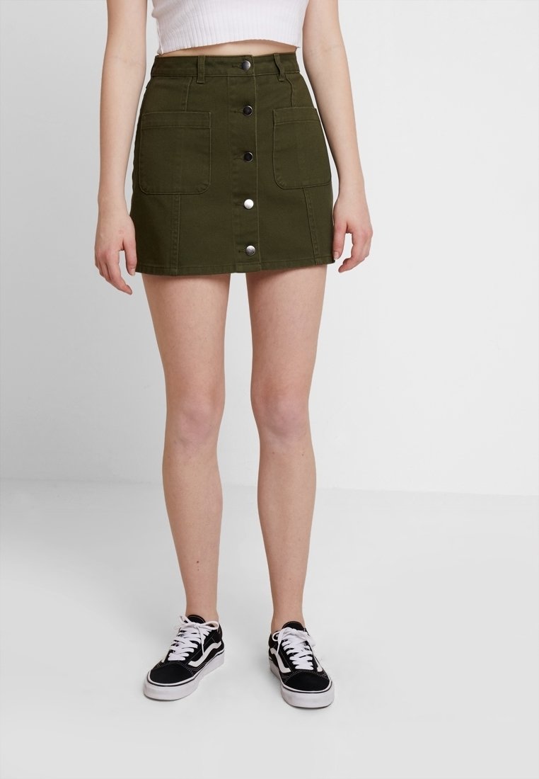 Femme portant une minijupe boutonnée devant vert olive avec deux poches avant et des baskets noires et blanches.