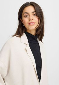 Béžový oversize blazer s výstrihom do "V", ľahký materiál a otvorený predný diel. Nosený cez čierny rolák. Hladká textúra.