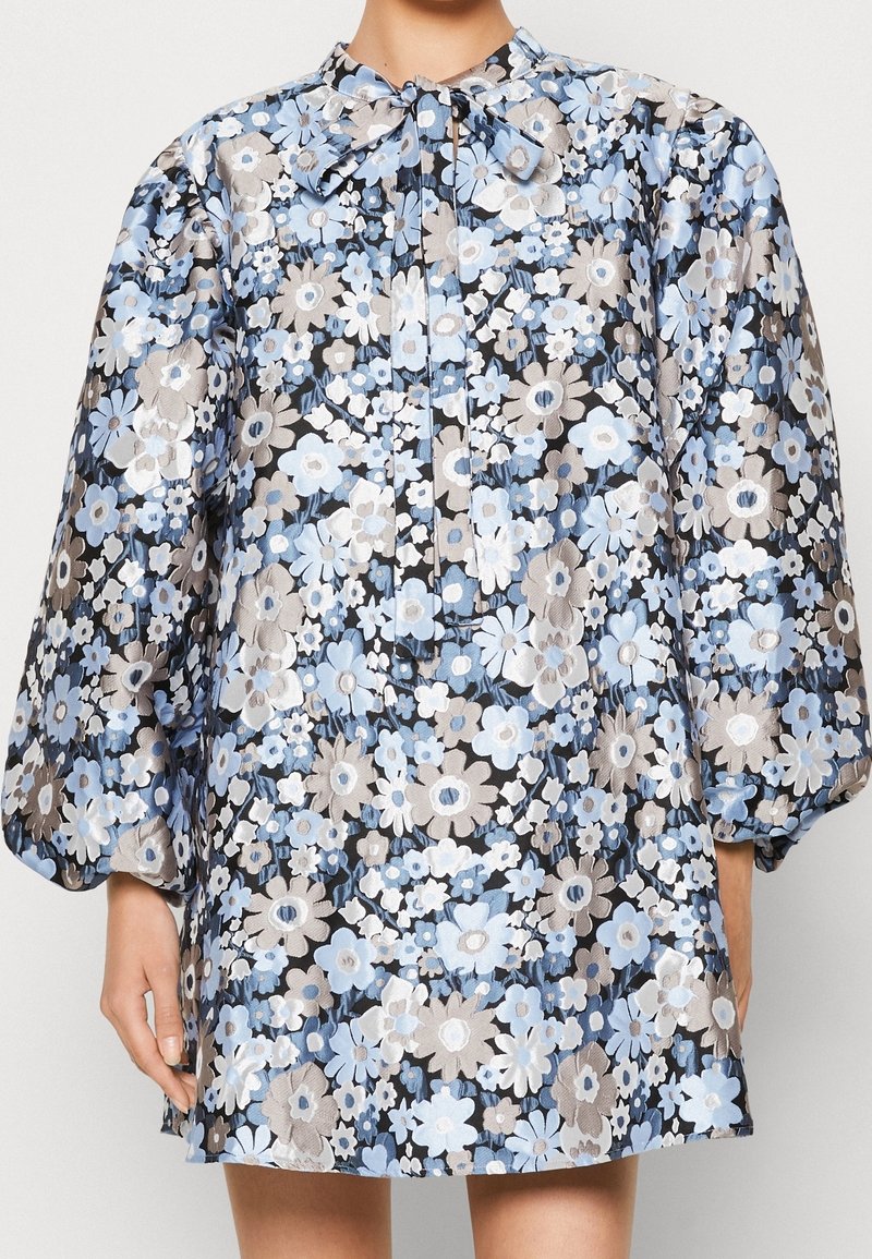 Robe en brocart floral avec des fleurs bleues, blanches et beiges, manches longues bouffantes et un nœud papillon assorti en tissu au col.