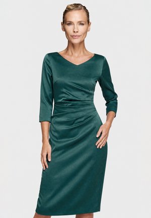 Woman with blonde hair wearing a fitted, dark green satin dress with three-quarter sleeves and a V-neckline, standing against a plain gray background.