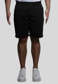 Pantaloni corti neri con taglio dritto, orlo risvoltato, tasche frontali e chiusura a un bottone, abbinati a sneakers bianche.