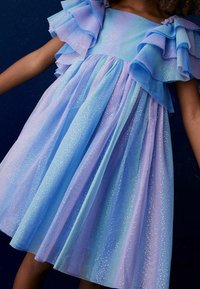 Hihaton mekko, jossa on kerroksittain aaltoilevia röyhelöitä sinisen ja laventelin liukuvärjäyksellä, koristeltu kimaltavalla tüllikankaalla ja vartalonmyötäisellä yläosalla.