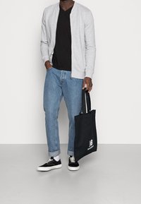 Chaqueta bomber gris con cremallera, camiseta negra de cuello en V, jeans azul claro, zapatillas negras, sosteniendo una bolsa tote negra con un logo blanco.