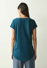 Tealfärgad topp med kort ärm och rund nederkant, tillverkad av ett mjukt material. Baksidan har en enkel design och en avslappnad passform.