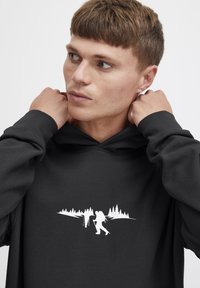 Joven con cabello corto castaño ajustándose una sudadera negra con capucha que presenta un gráfico blanco de dos excursionistas y árboles de pino en el pecho.