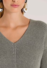 Grauer Strickpullover mit V-Ausschnitt und gerippter Textur, ergänzt durch große schwarz-weiße Ohrringe mit einem goldenen Akzent.