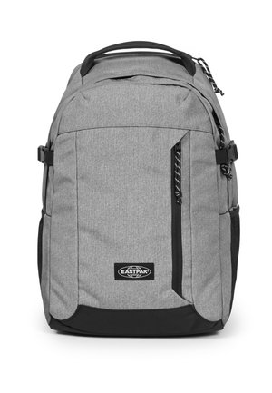 Mochila de tela gris con un compartimento principal y un bolsillo frontal con cremallera. Presenta acentos negros, un parche con el logo y asas de transporte. Superficie texturizada.