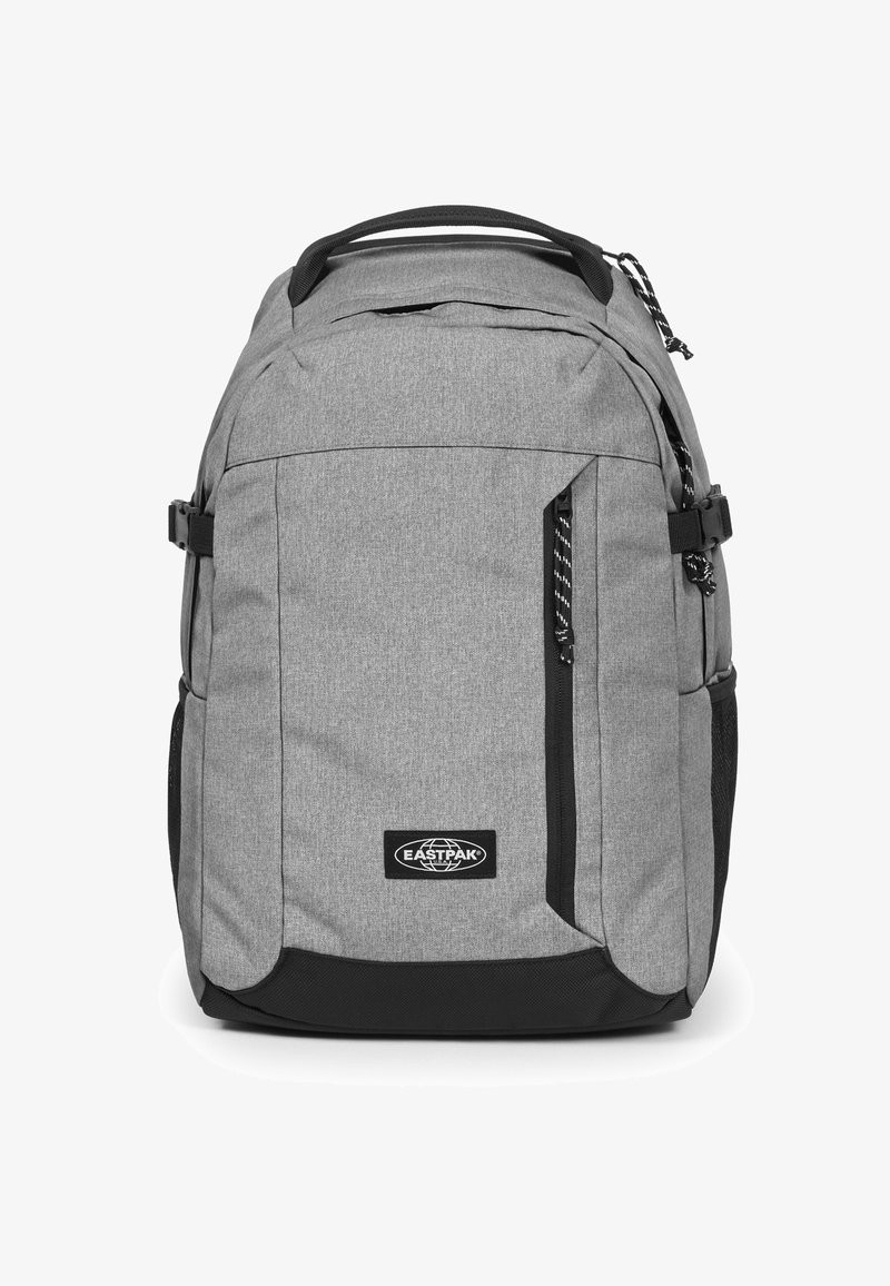 Mochila de tela gris con un compartimento principal y un bolsillo frontal con cremallera. Presenta acentos negros, un parche con el logo y asas de transporte. Superficie texturizada.