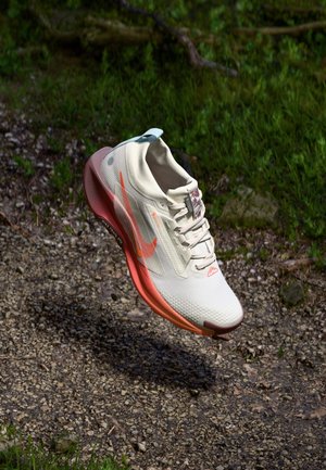 Lightweight, cream running shoe with orange accents, textured mesh upper, cushioned pink sole, and a blue heel tab, floating above a gravel surface.