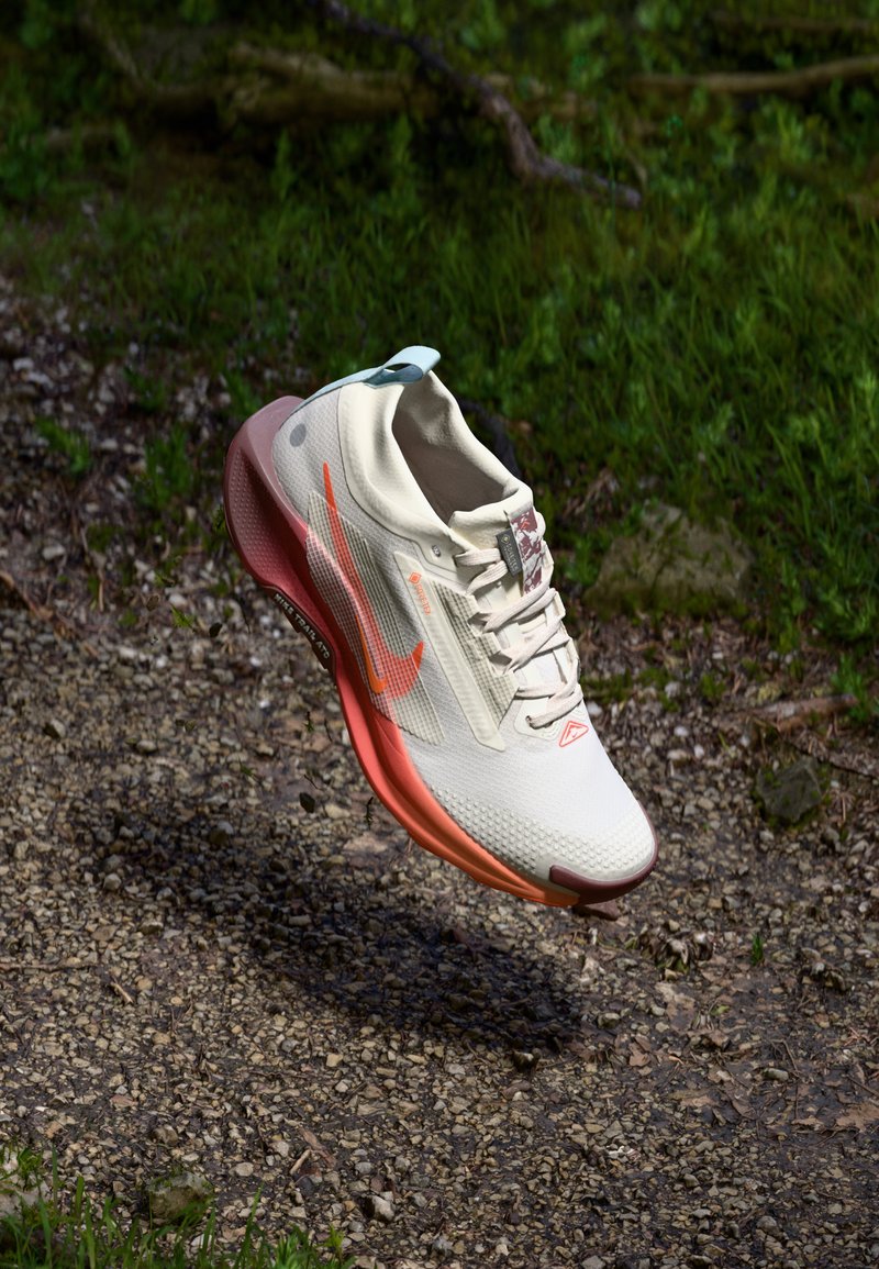 Chaussure de course légère, en crème avec des accents orange, tige en maille texturée, semelle rose rembourrée et onglet de talon bleu, flottant au-dessus d'une surface de gravier.