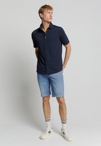 Marineblauw overhemd met korte mouwen en een knoopsluiting aan de voorkant, in combinatie met lichtblauwe denimshorts en witte sneakers met marineblauwe accenten en gestreepte sokken.