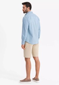 Camicia a maniche lunghe blu chiaro con polsini a bottone, abbinata a pantaloni corti beige e scarpe slip-on marroni. Design semplice, senza pattern visibili.