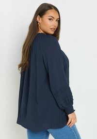 Blusa blu navy a maniche lunghe con texture plissettata, dettagli in pizzo sui polsini, vestibilità rilassata e scollatura rotonda, abbinata a jeans blu.