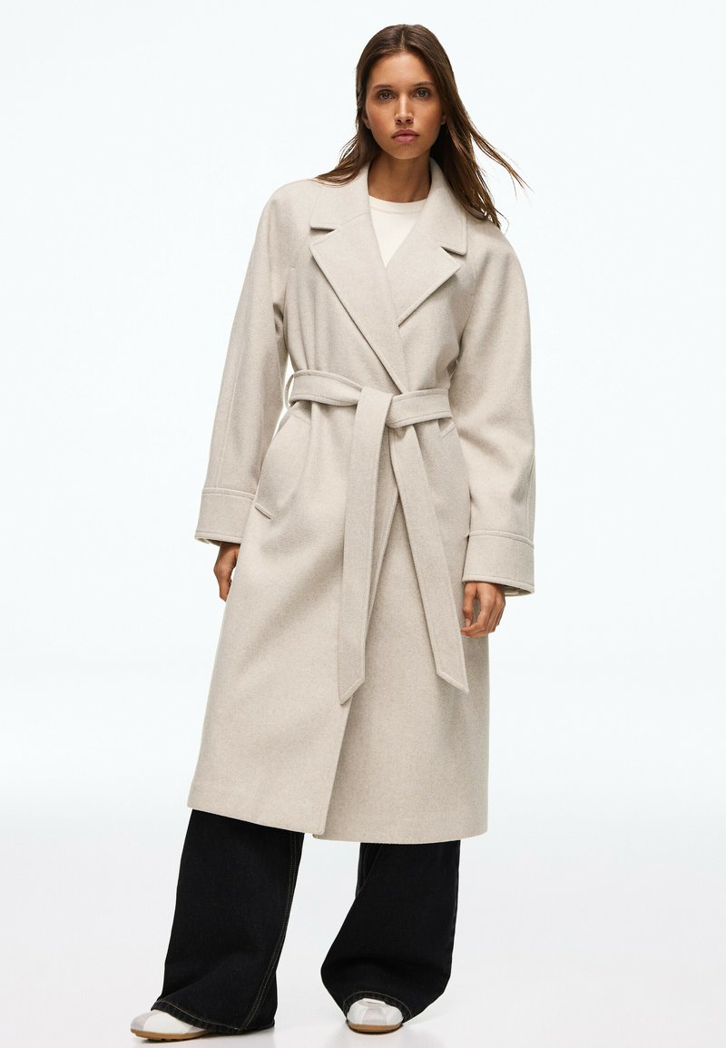 Femme portant un long manteau beige ceinturé avec un pantalon noir à jambes larges et des chaussures blanches, debout devant un fond clair uni.