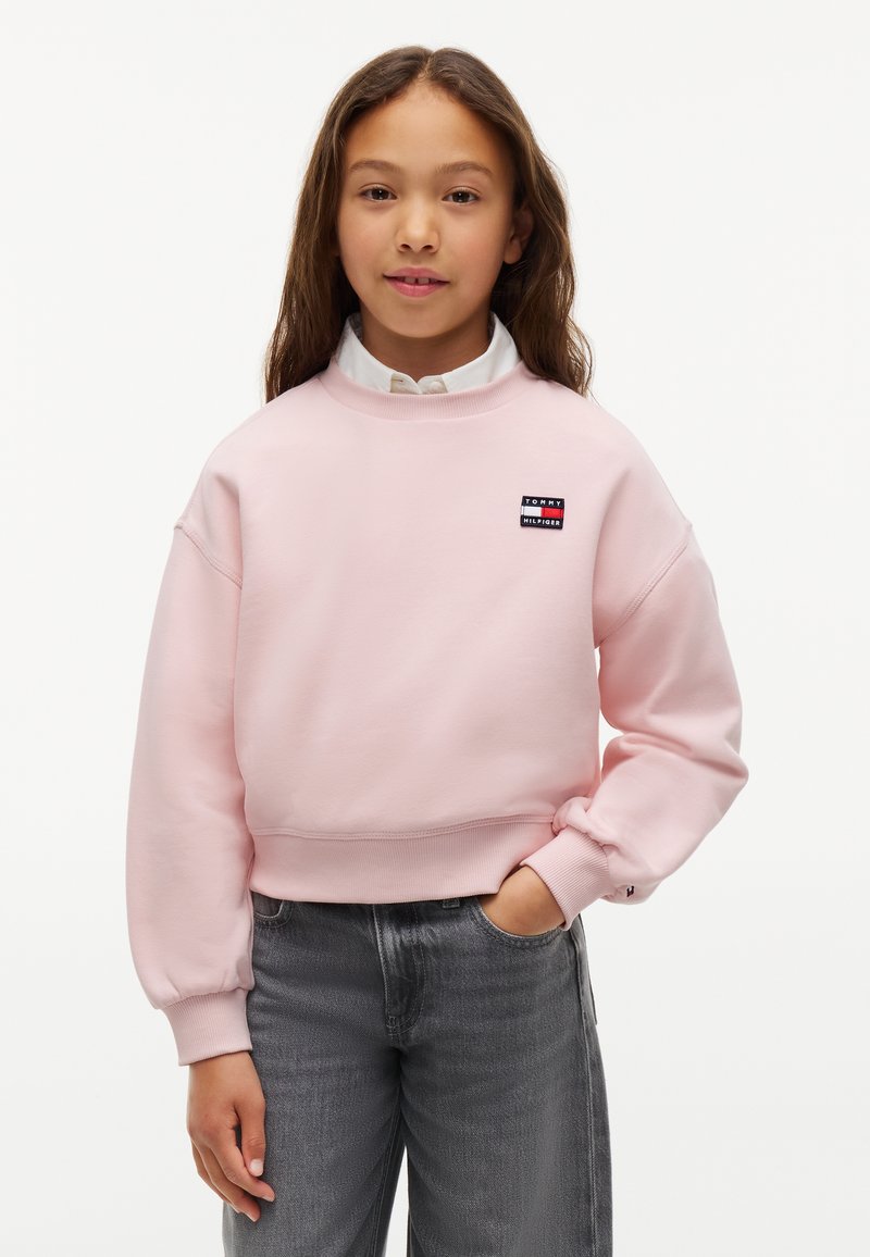 Fille aux longs cheveux bruns porte un sweat-shirt rose clair Tommy Hilfiger par-dessus une chemise blanche et un jean gris, sa main droite dans la poche.