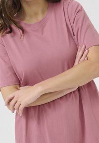 T-shirt en coton rose clair avec manches courtes et encolure ronde. Le tissu semble doux avec une texture lisse et des détails de couture minimaux.