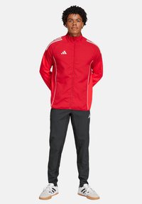 Rode sportieve jas met witte accenten en rits, gecombineerd met zwarte broeken en witte sportschoenen met zwarte strepen.