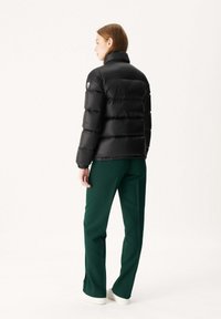 Zwarte pufferjas met een hoge kraag en horizontale stiksels; gecombineerd met groene broeken en witte sneakers. Logo-opdruk op de mouw.
