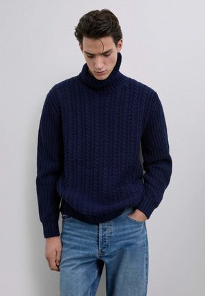 Suéter azul marino de punto trenzado con cuello alto y una tela gruesa y texturizada. Presenta mangas largas y un ajuste relajado, combinado con jeans azul claro.