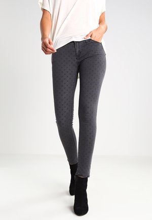 Personne portant un jean skinny gris foncé avec un motif à pois subtil, un haut blanc et des bottines noires, debout avec une main dans la poche.
