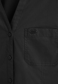 Svart knappad skjorta med V-hals, slät textur och en bröstficka med dekorativ accent. Knapparna är också svarta.