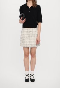 Polo negra de manga corta, minifalda de tweed blanca con flecos, zapatos negros con tiras y calcetines blancos hasta el tobillo. Bolso de mano sujeto con la mano.