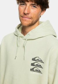 Sudadera de color verde claro con un bolsillo canguro, capucha con cordón y un gráfico negro de montaña y ola en el pecho. Tejido suave y casual.